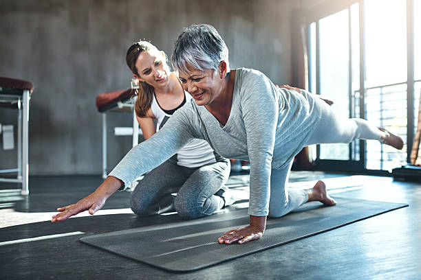 Senior woman doing a balance exercise on a yoga mat with guidance from a female fitness trainer.