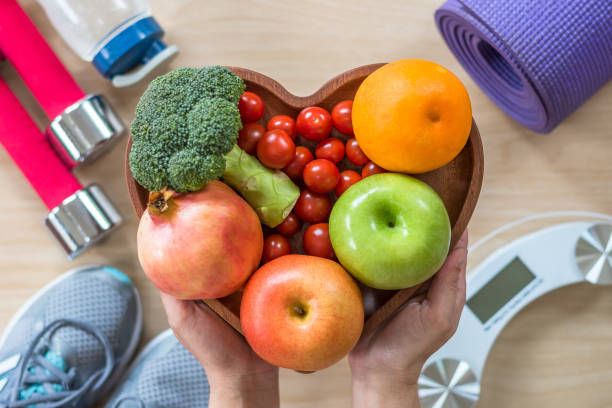 Fresh fruits and vegetables in a heart-shaped bowl surrounded by fitness equipment.