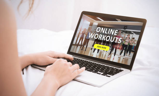 A woman using a laptop to access Herburn Studio’s online ladies gym workouts, featuring a fitness class with women exercising together on screen.WORKOUTS" and a "JOIN NOW" button displayed on the screen.